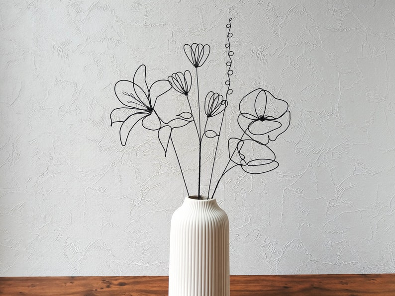 Puede incluir: Un jarr&oacute;n de cer&aacute;mica blanco con nervaduras verticales contiene un ramo de flores de alambre negro. Las flores son representaciones estilizadas de lirios, amapolas y otras flores.