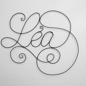 May include: A wire sculpture of the name "Lea" in a cursive font. The sculpture is made of black wire and is hanging on a white wall.