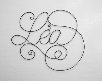 Puede incluir: Una escultura de alambre del nombre "Lea" en una fuente cursiva. La escultura está hecha de alambre negro y está colgada en una pared blanca.