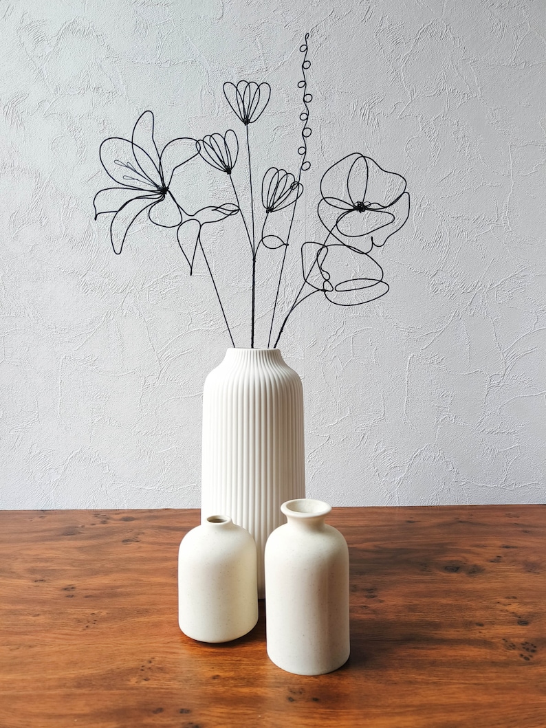 Puede incluir: Tres jarrones de cer&aacute;mica blanca, uno alto y dos m&aacute;s bajos, con una escultura de flores de alambre negro en el jarr&oacute;n alto. Los jarrones est&aacute;n sobre una mesa de madera.