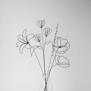 Puede incluir: Un ramo de flores de alambre negro en un jarr&oacute;n de vidrio transparente. Las flores son estilizadas e incluyen un lirio, una amapola y una flor m&aacute;s peque&ntilde;a con forma redondeada.