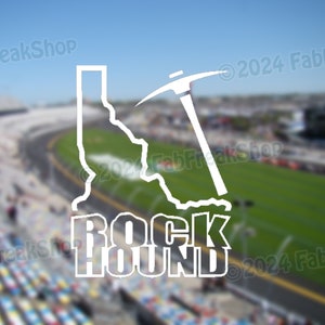 May include: White vinyl decal of the state of North Carolina with a pickaxe and the words "Rock Hound" underneath. The decal is on a blurred background of a race track.