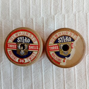 May include: Two vintage wooden thread spools with circular paper labels. The labels feature red, white, and blue graphics with the text DEWHURST'S SYLKO THREE SHELLS MACHINE TWIST 36. The paper labels show signs of wear, including tears and holes.