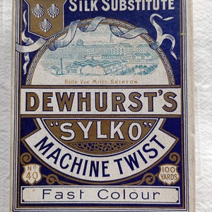May include: A vintage box of Dewhurst's Sylko Machine Twist thread. The box is blue and gold, with the text "Silk Substitute" and "Fast Colour." It features an illustration of a building and the text "Belle Vue Mills, Skipton." The box contains 91.44 metres of thread.