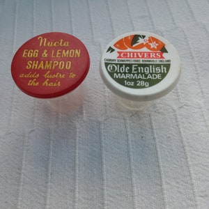 May include: Two small, round, plastic containers with lids. The red lid has gold lettering that reads "Nucta Egg & Lemon Shampoo adds lustre to the hair". The white lid has gold lettering that reads "Chivers Cadbury Schweppes Foods Bournville England Olde English Marmalade 1 oz 28g".