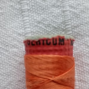 May include: Close-up of an orange thread spool with a red top. The word "COTTON" is printed on the top. The thread is tightly wound, creating a textured look. The background is a white, textured fabric, likely a towel.