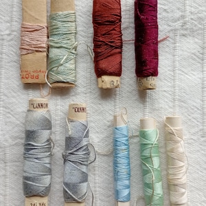 May include: A selection of vintage thread spools in shades of pink, green, red, and blue. The spools are made of cardboard and paper, some labelled "CANNON" and "LISTER". The thread is likely rayon.