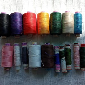 May include: A selection of sewing threads in various colours, including black, purple, red, yellow, green, beige, and blue. Smaller spools in pink, white, navy, and brown are also visible. The threads are arranged on a white textured surface.