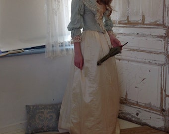 May include: A woman in a light blue and white dress with a lace bodice and a white skirt. She is holding a small, green parasol in her right hand. She is standing in front of a white door with a white lace curtain behind her.