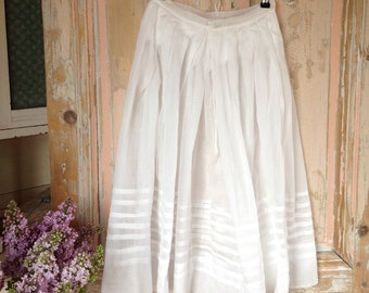 May include: A white, semi-sheer skirt with horizontal stripes near the hem, hanging on a metal hanger. The skirt is pleated and appears to be made of a lightweight fabric. The background is a weathered, light-colored wooden door.