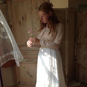 May include: A long, white, vintage-style dress with lace trim and a sheer, cream-colored top. The dress is worn in a sunlit room with a wooden door and a vase of pink flowers. The dress appears to be made of a lightweight fabric.