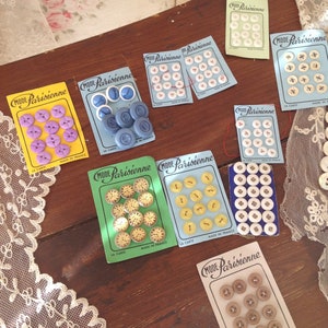 Op de afbeelding: Assortiment van kleurrijke knopen op kaarten met de tekst "Mode Parisienne" en "La Carte" in het Frans. De knopen zijn gemaakt in Frankrijk en zijn verkrijgbaar in verschillende kleuren, waaronder blauw, geel, wit en bruin.