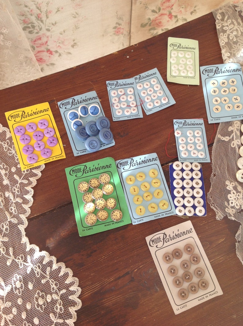 Op de afbeelding: Assortiment van kleurrijke knopen op kaarten met de tekst "Mode Parisienne" en "La Carte" in het Frans. De knopen zijn gemaakt in Frankrijk en zijn verkrijgbaar in verschillende kleuren, waaronder blauw, geel, wit en bruin.