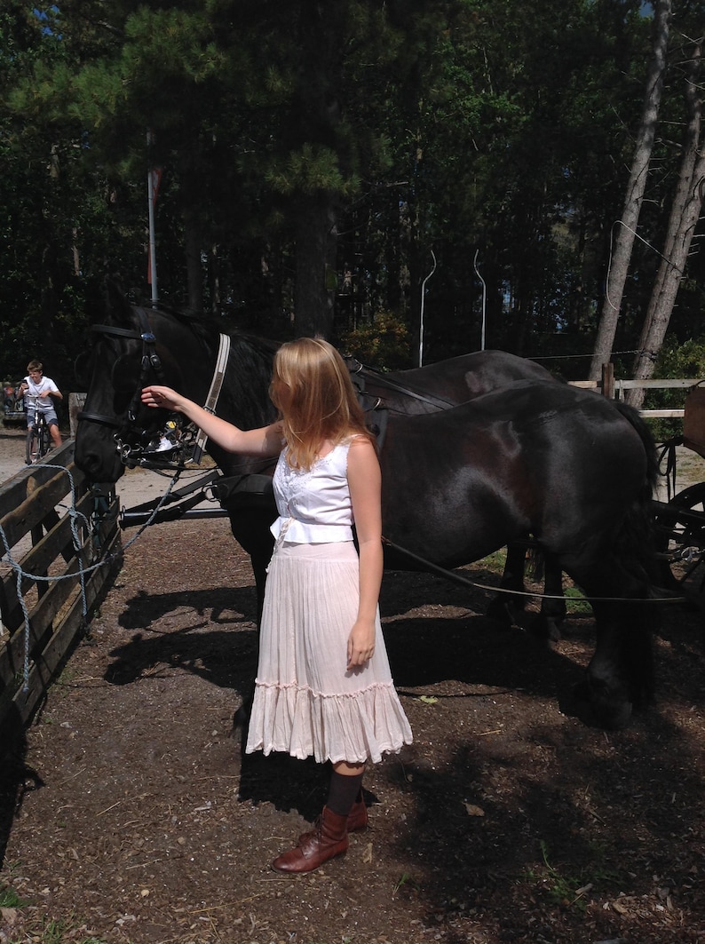 May include: A woman stands near a black horse harnessed to a carriage. She wears a white top, a light pink skirt, and brown boots. The horse is in front of a wooden fence, with trees in the background.