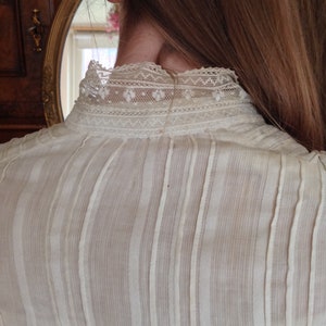 Puede incluir: Una blusa blanca, transparente, con rayas verticales y un delicado cuello de encaje. La blusa est&aacute; hecha de una tela ligera y tiene un corte holgado.