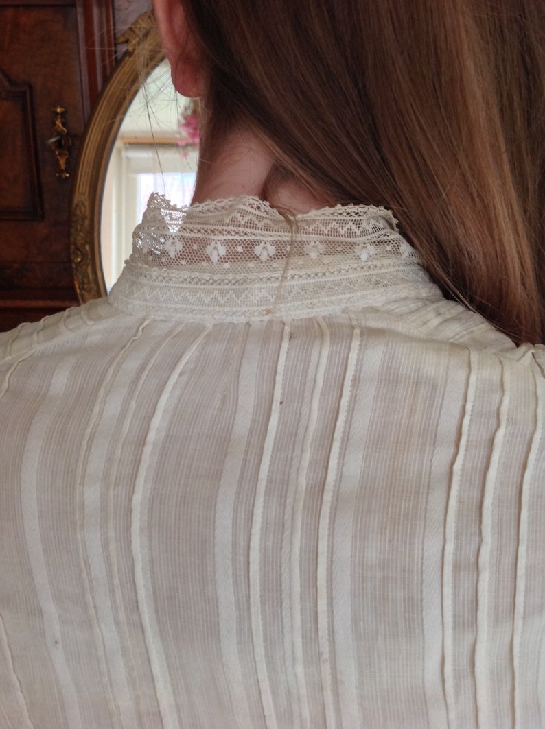 Puede incluir: Una blusa blanca, transparente, con rayas verticales y un delicado cuello de encaje. La blusa est&aacute; hecha de una tela ligera y tiene un corte holgado.