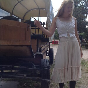 May include: A wooden covered wagon with a canvas top. The wagon has black wheels and is parked on a gravel surface. A person is standing next to the wagon, wearing a white top and a long, cream-colored skirt.