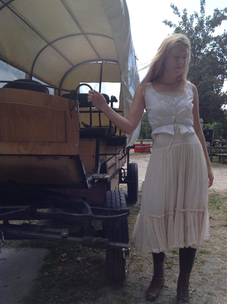 May include: A wooden covered wagon with a canvas top. The wagon has black wheels and is parked on a gravel surface. A person is standing next to the wagon, wearing a white top and a long, cream-colored skirt.