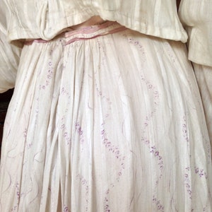 Puede incluir: Un vestido de algod&oacute;n blanco con una cinta de raso rosa y un delicado estampado floral en morado.