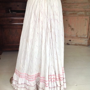 Puede incluir: Una falda de algod&oacute;n blanca con un delicado estampado floral y ribete de cinta rosa. La falda tiene varias capas de volantes en el dobladillo.