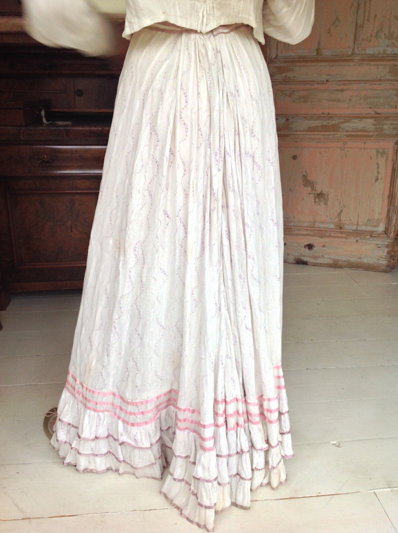 Puede incluir: Una falda de algod&oacute;n blanca con un delicado estampado floral y ribete de cinta rosa. La falda tiene varias capas de volantes en el dobladillo.