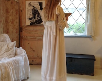 May include: A floor-length, long-sleeved cream-coloured dress with a high waistline, a gathered skirt, and horizontal bands of sheer fabric near the hem. The dress features tie-style shoulder details and a simple, flowing silhouette.