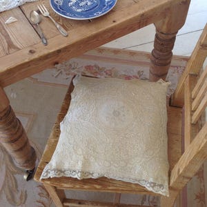 Könnte beinhalten: Ein quadratischer, cremefarbener Kissenbezug mit aufwendigen Spitzendetails und einem zentralen, gestickten Blumenmotiv. Das Textil liegt auf der Sitzfläche eines Holzstuhls und zeigt zarte, gewellte Ränder sowie eine feine Webstruktur.