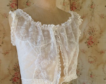 May include: A white lace top with a ruffled neckline and a tie at the front. The top is on a dress form and is against a floral patterned background.