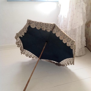 Op de afbeelding: Een parasol in vintage-stijl met een donkerblauwe luifel en een decoratieve kanten rand. De parasol heeft een houten handvat en is open, waardoor het ingewikkelde ontwerp zichtbaar is. De kanten rand voegt een delicate touch toe.