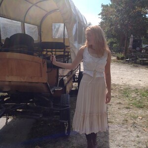 May include: A wooden covered wagon with a canvas top. The wagon is brown and tan, with black wheels. A person is standing next to the wagon, wearing a white top and a long, cream-colored skirt.