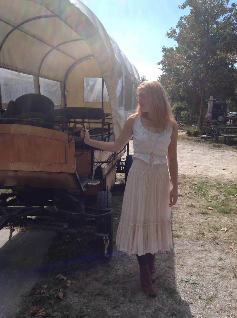 May include: A wooden covered wagon with a canvas top. The wagon is brown and tan, with black wheels. A person is standing next to the wagon, wearing a white top and a long, cream-colored skirt.