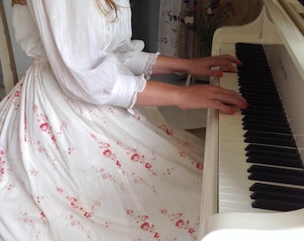 May include: A white long-sleeved dress featuring a delicate red floral print and a wide lace hem, worn while playing a white grand piano. The dress has a high neckline and gathered fabric at the waist, with lace detailing on the sleeve cuffs.