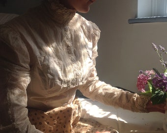 May include: A woman wearing a white lace blouse with a high collar and a floral patterned skirt sits on a bed, holding a bouquet of purple flowers.