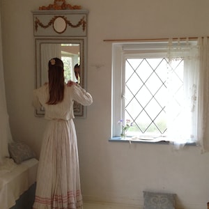 Puede incluir: Una mujer con un vestido blanco con ribetes rosas se para frente a un espejo en una habitaci&oacute;n con una ventana. El espejo tiene un marco adornado y un dise&ntilde;o floral. La ventana tiene un panel de forma de diamante y cortinas blancas.