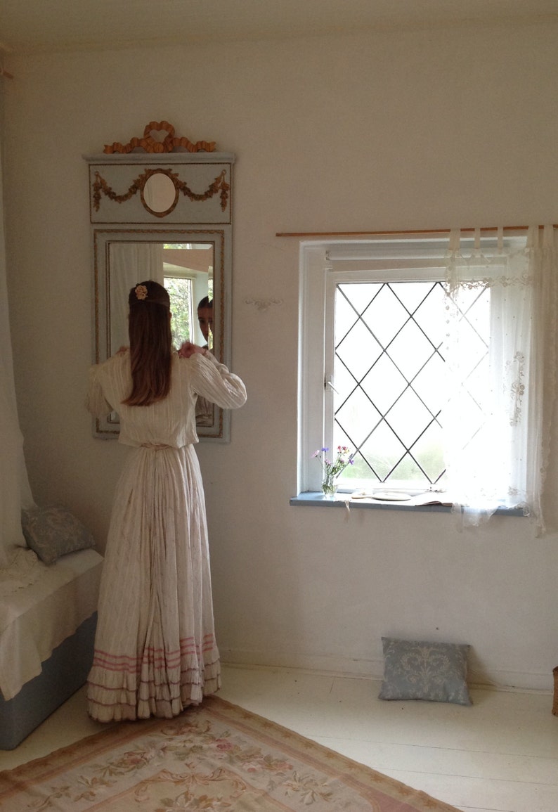 Puede incluir: Una mujer con un vestido blanco con ribetes rosas se para frente a un espejo en una habitaci&oacute;n con una ventana. El espejo tiene un marco adornado y un dise&ntilde;o floral. La ventana tiene un panel de forma de diamante y cortinas blancas.