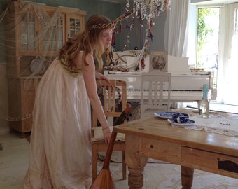 Puede incluir: Una mujer con un vestido blanco y una corona en la cabeza se encuentra junto a una mesa de madera y un piano blanco. Está sosteniendo un laúd de madera. La habitación está decorada en un estilo vintage con paredes blancas y una lámpara de araña.