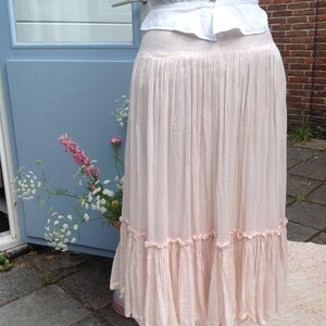 May include: A light pink, floor-length skirt with a ruffled hem. The skirt is paired with a white blouse. The skirt is displayed on a white mannequin. The background includes a blue door and brick wall.