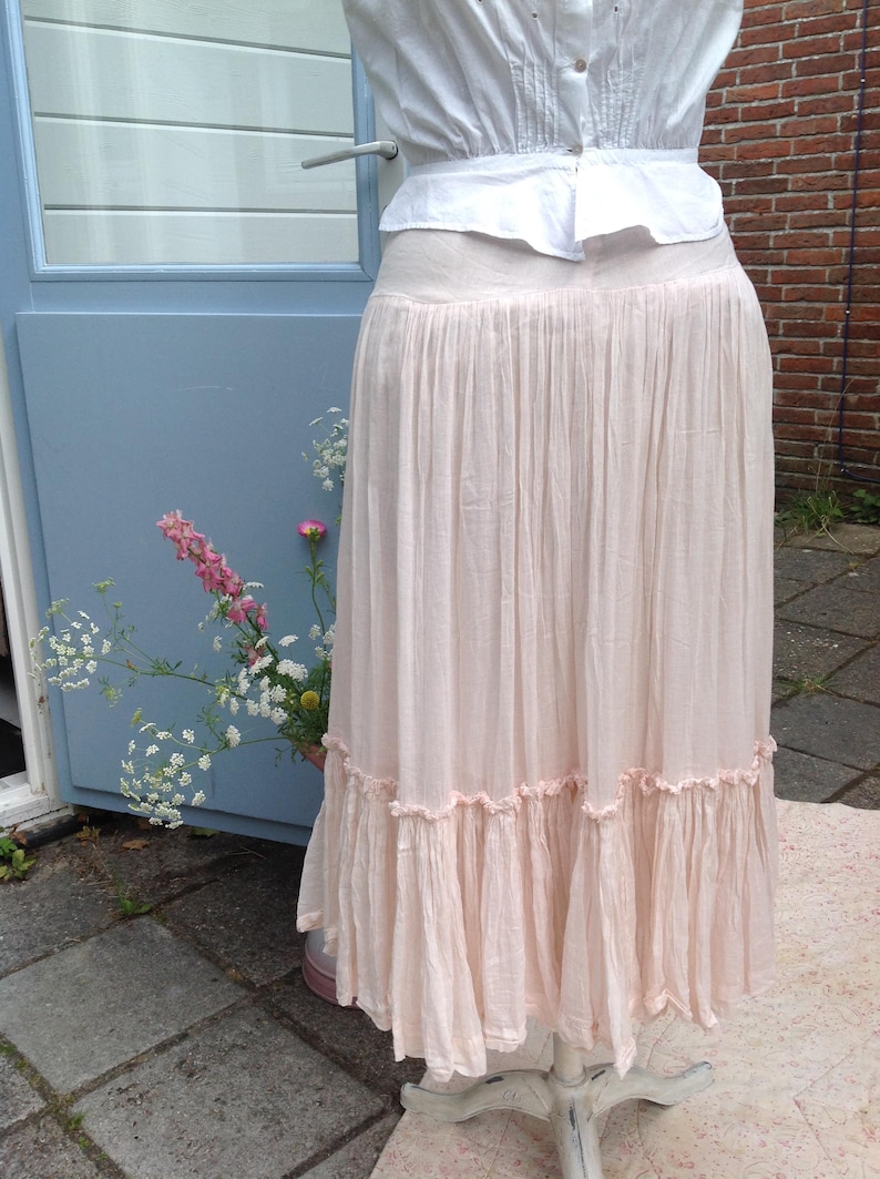 May include: A light pink, floor-length skirt with a ruffled hem. The skirt is paired with a white blouse. The skirt is displayed on a white mannequin. The background includes a blue door and brick wall.