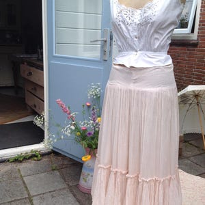 May include: A vintage-style outfit on a mannequin. The ensemble includes a white, embroidered top and a long, light pink skirt with ruffled trim. The outfit is displayed outdoors near a blue door and a vase of flowers.