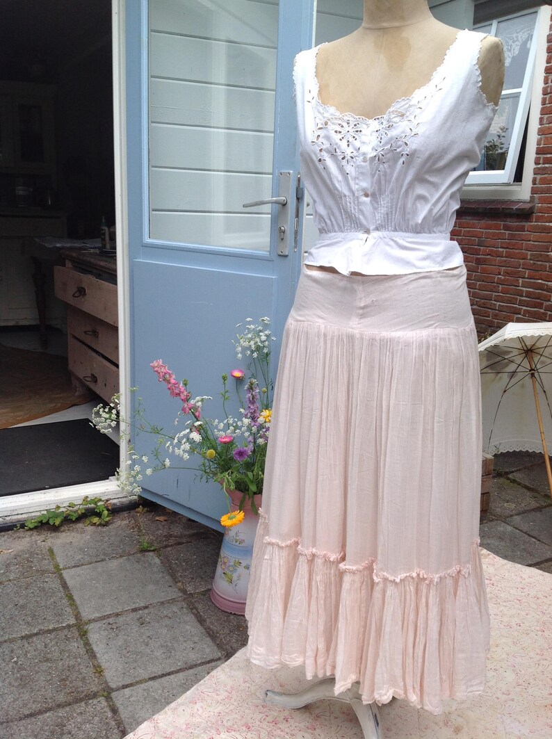 May include: A vintage-style outfit on a mannequin. The ensemble includes a white, embroidered top and a long, light pink skirt with ruffled trim. The outfit is displayed outdoors near a blue door and a vase of flowers.