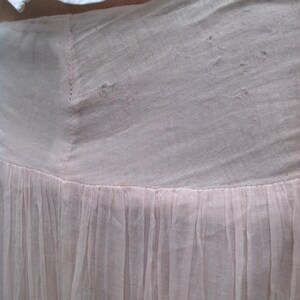 May include: Close-up of a light pink skirt with a gathered, flowing design. The skirt is made of a sheer fabric, with a fitted waist and a flared, pleated bottom. A white garment is visible at the top.