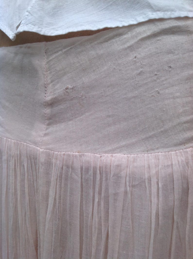 May include: Close-up of a light pink skirt with a gathered, flowing design. The skirt is made of a sheer fabric, with a fitted waist and a flared, pleated bottom. A white garment is visible at the top.