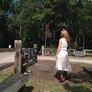 May include: A person wearing a white top and a light-colored skirt stands in a park-like setting. The person is facing away from the camera, with long hair. A wooden fence and bench are in the foreground.