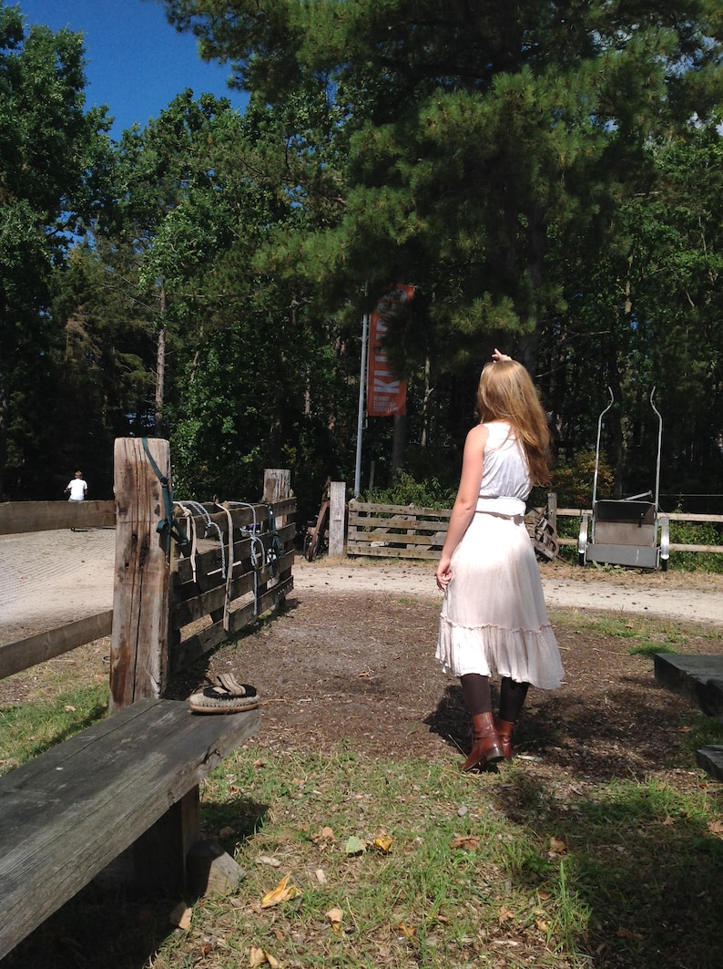 May include: A person wearing a white top and a light-colored skirt stands in a park-like setting. The person is facing away from the camera, with long hair. A wooden fence and bench are in the foreground.
