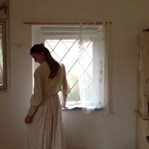 Puede incluir: Una mujer con un vestido blanco con ribetes rosas se encuentra en una habitaci&oacute;n con una ventana y un armario de madera. La ventana tiene una cortina de encaje blanco y una vista de un jard&iacute;n.