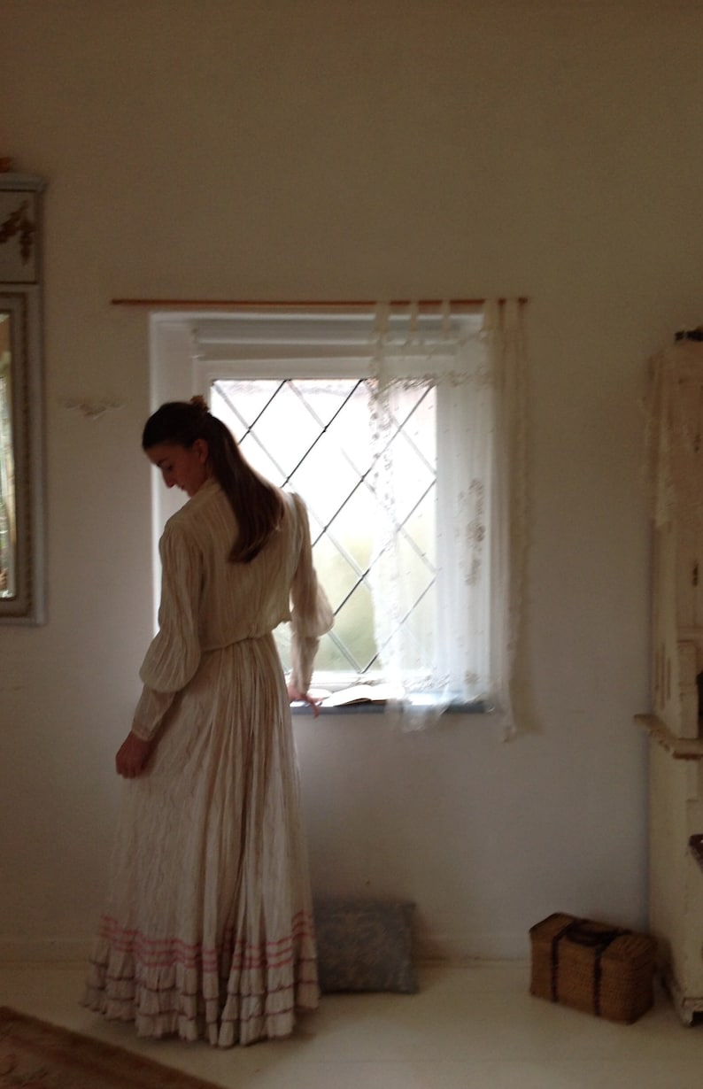 Puede incluir: Una mujer con un vestido blanco con ribetes rosas se encuentra en una habitaci&oacute;n con una ventana y un armario de madera. La ventana tiene una cortina de encaje blanco y una vista de un jard&iacute;n.