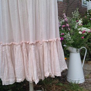 May include: A light pink, ruffled skirt displayed on a white, distressed pedestal. The skirt has a layered design with a ruffled hem. A white pitcher filled with flowers sits nearby in a garden setting.