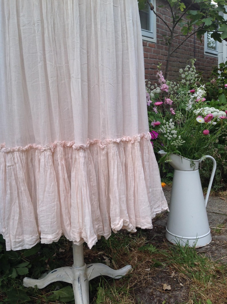 May include: A light pink, ruffled skirt displayed on a white, distressed pedestal. The skirt has a layered design with a ruffled hem. A white pitcher filled with flowers sits nearby in a garden setting.