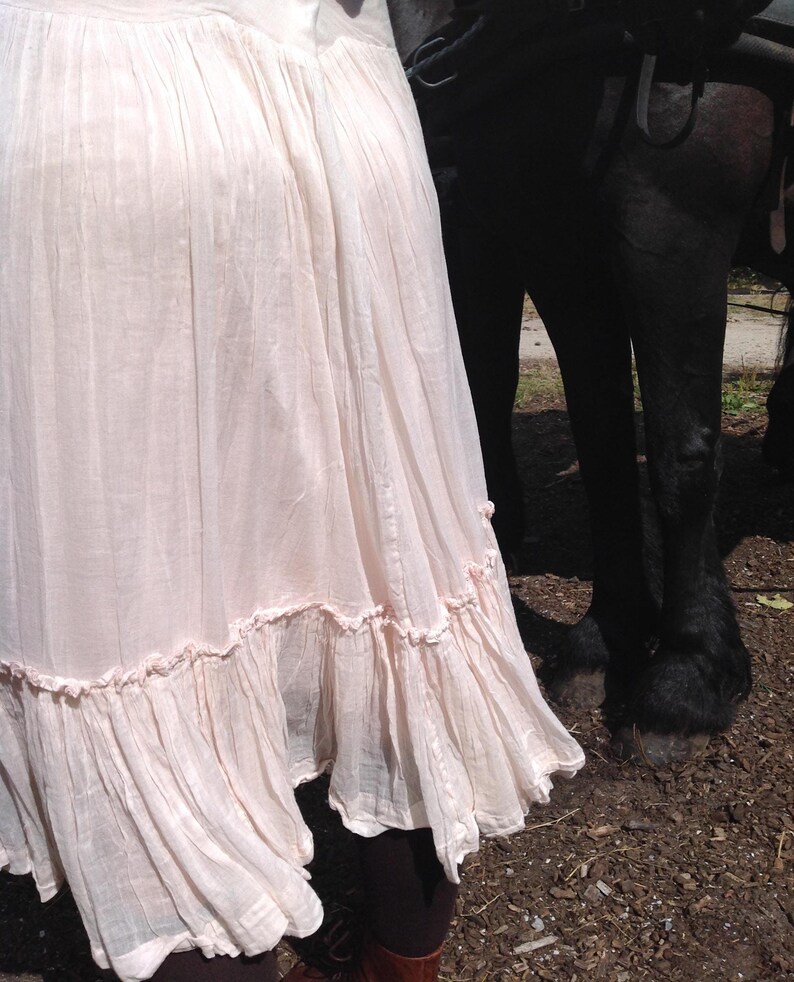 May include: A light pink, flowing skirt with a ruffled hem. The skirt is made of a lightweight fabric and has a soft, textured appearance. The skirt is part of a bohemian style outfit.