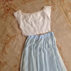 Puede incluir: Un top corto blanco con un escote ancho y un delicado borde de encaje a lo largo del cuello, combinado con una falda plisada de color azul claro con una cinturilla sólida. El conjunto consta de dos piezas separadas, una blusa sin mangas y una falda fluida.